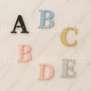 May include: Five colorful uppercase letters, A, B, C, D, and E, are arranged on a light-colored surface. The letters are in black, light blue, gold, pink, and silver. The letters are made of a solid material.