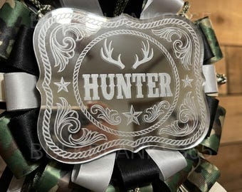 May include: A decorative badge with the word "HUNTER" in a Western-style font, featuring deer antlers and star accents. The badge is surrounded by a decorative border and set against a backdrop of camouflage, black, and silver ribbons.