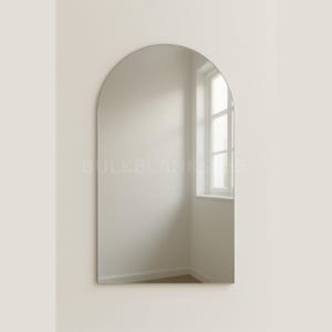 May include: A large, frameless, arched mirror. The mirror is mounted on a light beige wall. The reflection shows a window and a room with white walls and a wooden floor. The mirror's shape is a rounded arch.
