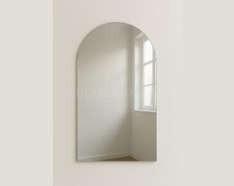 May include: A large, frameless, arched mirror. The mirror is mounted on a light beige wall. The reflection shows a window and a room with white walls and a wooden floor. The mirror's shape is a rounded arch.