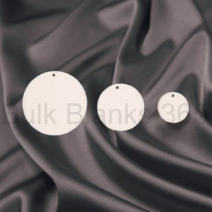 May include: Three white ceramic round ornaments with a hole at the top. The ornaments are different sizes and are arranged on a dark gray fabric background.