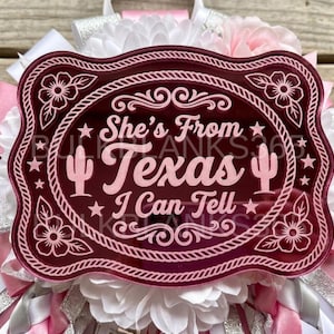 May include: A maroon acrylic plaque with a rope-style border and floral corners, featuring the light pink text "She's From Texas I Can Tell" and two cactus icons. The plaque is mounted on a decorative base of white and pink fabric ribbons.