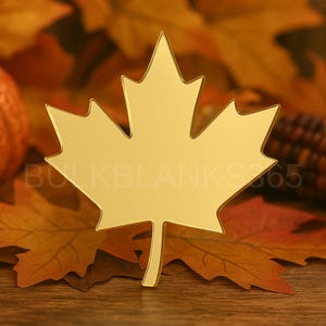 May include: A gold-colored maple leaf-shaped object, possibly a blank for crafting or decoration, is the focal point. It rests on a bed of autumn leaves in shades of orange and brown. The text "BULKBLANKS365" is visible.