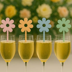 May include: Four champagne flutes filled with a golden liquid, each adorned with a colorful flower-shaped drink stirrer. The stirrers are pastel pink, orange, blue, and green. A blurred floral arrangement is in the background.
