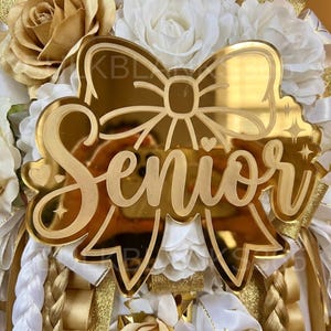 May include: A gold-colored acrylic bow-shaped decoration with the word "Senior" in a cursive font. The decoration is surrounded by white and gold flowers and ribbons, creating a celebratory theme.