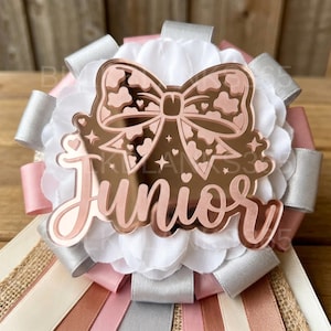 May include: A decorative rosette featuring a layered white fabric flower base, accented with silver and pink satin ribbons and burlap strips. Centered is a rose gold mirrored acrylic topper shaped like a bow with the word Junior in script.