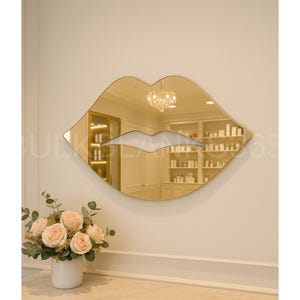May include: A gold-toned lip-shaped mirror hangs on a cream-colored wall. The mirror reflects a room with shelves and a chandelier. A bouquet of pink roses in a white vase sits below the mirror.