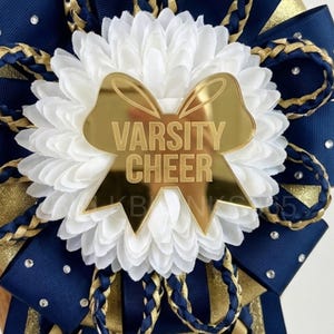May include: A cheerleading mum with a gold-colored bow that reads "VARSITY CHEER". The mum features a white flower center, navy blue and gold ribbons, and decorative rhinestones. Braided gold and navy blue accents complete the design.
