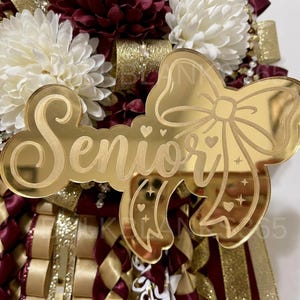 May include: A gold mirror acrylic cutout featuring the word Senior in cursive script, integrated with a large bow design. The bow is decorated with small etched hearts and four-pointed stars. It is layered over a background of white and maroon flowers.
