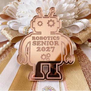 May include: A rose gold mirrored acrylic robot cutout featuring the text ROBOTICS SENIOR 2027 and gear icons, centered on a decorative rosette made of white and gold ribbons, pearl strands, and small clear rhinestones.