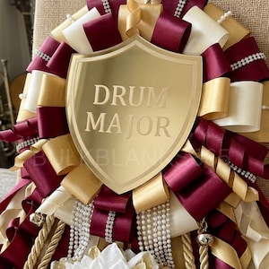 May include: A decorative gold shield with the words "DRUM MAJOR" is the centerpiece of a large ribbon arrangement. The ribbons are in burgundy, gold, and cream colors, accented with pearl and rhinestone strands. A bell and rope add to the design.