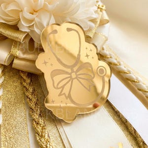 May include: A gold-colored stethoscope-shaped badge with a bow and star accents. The badge is set against a backdrop of white and gold ribbons and a cream-colored flower, creating a celebratory theme.