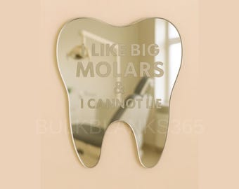 May include: A tooth-shaped mirror with the text "I LIKE BIG MOLARS & I CANNOT LIE" etched into the reflective surface. The mirror is mounted on a light peach-colored wall, reflecting a dental office.