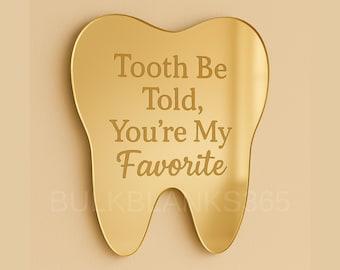 May include: A gold-colored, tooth-shaped plaque with the engraved text "Tooth Be Told, You're My Favorite." The plaque has a smooth, reflective surface and is mounted on a light beige background.