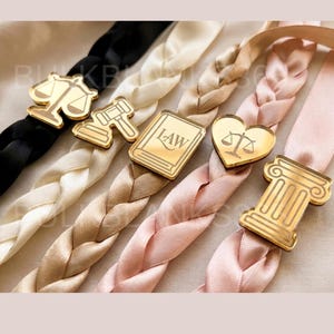 May include: Close-up of several braided ribbons in black, cream, tan, and pink, each adorned with a gold-colored charm. The charms feature symbols like scales, a gavel, a book with "LAW" text, a heart with scales, and a pillar.
