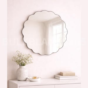 May include: A round, scalloped-edge mirror with a black frame hangs on a white wall. Below the mirror, a white vase holds white flowers. A small white bowl and stacked books sit on a white dresser.