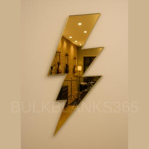 May include: A gold-colored, lightning bolt-shaped mirror mounted on a beige wall. The mirror reflects a room with musical instruments and a brick wall. The text "BULKBLANKS365" is visible at the bottom.