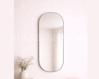 May include: An oval-shaped mirror with a black frame. The mirror is tall and narrow, reflecting a bright room. A white vase with flowers sits below the mirror. The wall is a light pink color.