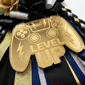 May include: A gold-colored acrylic charm in the shape of a video game controller with the words "LEVEL UP". The charm is decorated with lightning bolts and is set against a backdrop of black, gold, silver, and blue ribbons.