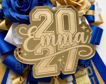 May include: A gold mirrored acrylic topper featuring the name Emma and the year 2024 in a layered design with star accents. The topper is set against a background of blue and gold fabric ribbons and artificial roses in blue and gold tones.