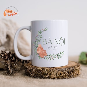 May include: White ceramic mug with a green wreath design and the text "BÁ NÕI est 24". The wreath includes a small pink hat and booties.