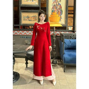 May include: A red long-sleeved dress with a white floral design on the chest. The dress has a boat neck and a side slit. The model is wearing white shoes.