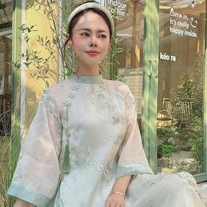 May include: A woman wearing a light green, floral patterned dress with a white headband and earrings. The dress has a mandarin collar and a tassel detail. The woman is standing in front of a green door with a sign that says "indoor coffee".