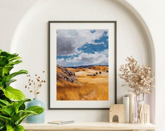 May include: Framed landscape art print depicting a rural scene with rolling hills, golden fields, and a cloudy blue sky. The artwork is displayed in a black frame, adding a touch of elegance to the naturalistic scene. The painting evokes a sense of tranquility.