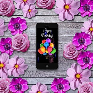May include: A black smartphone with a white screen displaying the text "Happy Birthday!" with colorful balloons and fireworks. The phone is surrounded by pink flowers on a wooden background.