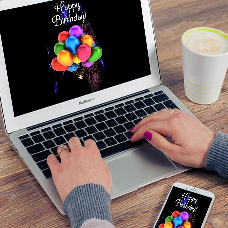 May include: A laptop computer with a black screen displaying a birthday greeting with colorful balloons and fireworks. The laptop is on a wooden table with a cup of coffee and a smartphone with a similar birthday greeting.
