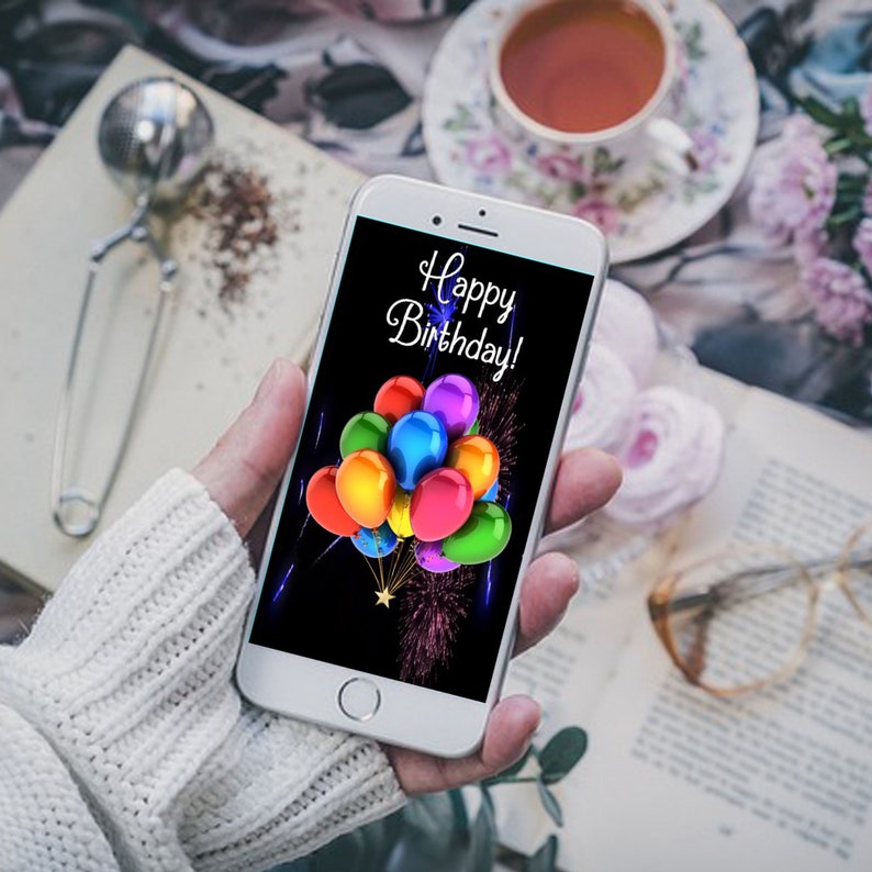 May include: A smartphone displaying a birthday greeting with colorful balloons and fireworks on a black background. The text on the screen reads "Happy Birthday!"