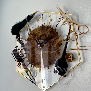 May include: A gold-framed hexagonal clock with a brown splatter design. The clock features black hairdressing tools, including scissors, a comb, a hair dryer, and a hair brush, as part of the design.