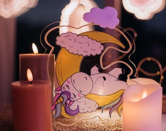 May include: A clear acrylic unicorn nightlight with a pink and yellow crescent moon and a cloud. The unicorn is sleeping on the moon. There are three lit candles in front of the nightlight.