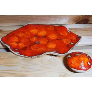 Könnte beinhalten: Keramik-Dekoration zum Thema Pilze. Das Bild zeigt eine große, blattförmige Schale und einen kleinen, runden Pilz, beide mit einem leuchtenden Rot- und Orangeton. Die Schale hat einen weißen Rand und orangefarbene Punkte.