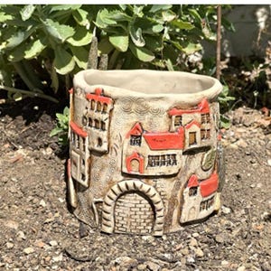 Könnte beinhalten: Beige Keramik-Blumentopf mit detailliertem Reliefdesign einer Dorflandschaft. Der Topf zeigt Gebäude mit roten Dächern, gewölbte Türen und strukturierte Wände, ideal für kleine Pflanzen oder Sukkulenten.