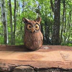 Könnte beinhalten: Eine bronzefarbene Eulenfigur mit großen, detaillierten Augen und einem strukturierten Körper. Die Eule sitzt auf einem Holzstück, mit einem verschwommenen Hintergrund eines Waldes. Die Figur ist etwa 10 cm hoch.