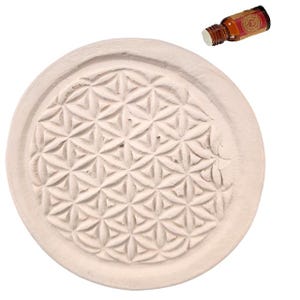 Könnte beinhalten: Ein kreisförmiger, cremefarbener Steinuntersetzer mit einem eingravierten geometrischen Blume des Lebens-Muster. Daneben steht eine kleine, bernsteinfarbene Glasflasche mit rotem Etikett und weißem Verschluss, die eine Flüssigkeit enthält.