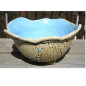 Könnte beinhalten: Handgefertigte Keramikschale mit hellblauem Innenraum und strukturierter, beigefarbener Außenseite. Die Schale weist geprägte Muster und kleine blaue Akzente auf. Der Rand hat ein wellenförmiges Design, das zu ihrem einzigartigen, künstlerischen Reiz beiträgt. Ein dekoratives Stück für das Zuhause.