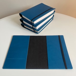 May include: A blue leather notebook cover with a black elastic closure. The cover is open to reveal a black interior. The cover is lying on a white surface.