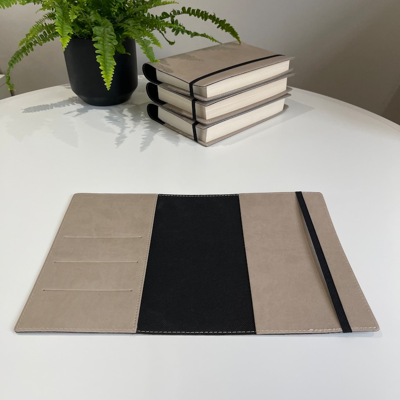 May include: A beige leather journal with a black elastic closure and a black leather interior. The journal has three pockets on the front cover.