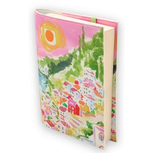 以下が含まれることがあります： ピンク、赤、白の建物、緑の丘、黄色の太陽が描かれた、水彩画の海岸沿いの町のカラフルな表紙の本。表紙はピンク色で、緑色のゴムバンドが付いています。本はブックバディです。
