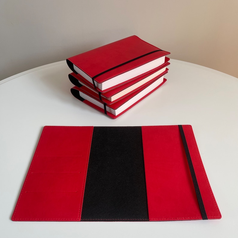May include: Red leather notebook cover with a black elastic closure. The cover is closed and lying flat on a white surface.