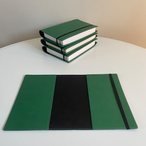 May include: A green leather notebook cover with a black elastic band closure. The cover is open to reveal a black interior. Three green leather notebook covers are stacked on a white surface.
