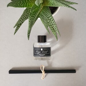 Op de afbeelding: Een heldere glazen fles met een zwarte dop en een zwart etiket met de tekst "Fuego Scent" en "Hand Made in Lithuania". De fles staat op een grijs oppervlak met zwarte diffuserstokken en een groene plant in een pot.