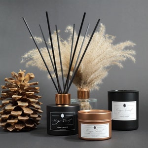 May include: Three black and gold scented candles with the text "Fragor Scent" and "Hand Made" on the labels. The candles are arranged on a gray surface with a pine cone and a vase of dried pampas grass.