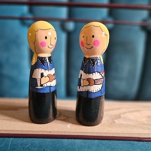 May include: Two wooden peg dolls with blonde hair, painted blue shirts, and holding guitars. The dolls are standing on a wooden surface.