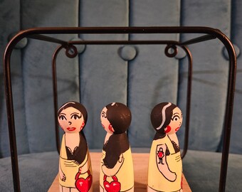 May include: Three wooden dolls with painted faces and black hair. Each doll is wearing a yellow dress with a red heart and is holding a red heart in their hand. The dolls are displayed in a black metal cube.