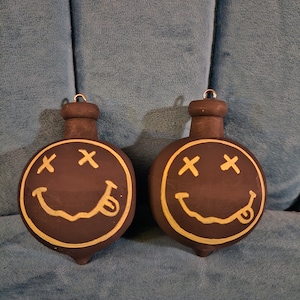 May include: Two black Christmas baubles shaped like a smiley face with a tongue sticking out. The eyes are marked with an 'X' and the face is outlined in yellow.