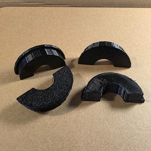 May include: Four black, semi-circular 3D-printed objects are arranged on a tan surface. The objects have a textured appearance and varying designs, with one appearing to be segmented. The objects are likely components or parts.
