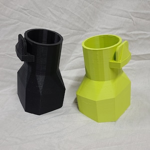 Puede incluir: Dos adaptadores impresos en 3D, uno negro y otro verde lima, cada uno con una sección superior cilíndrica, una base hexagonal más ancha y una pequeña pestaña que sobresale en el lateral. La textura de capas de la impresión es visible.